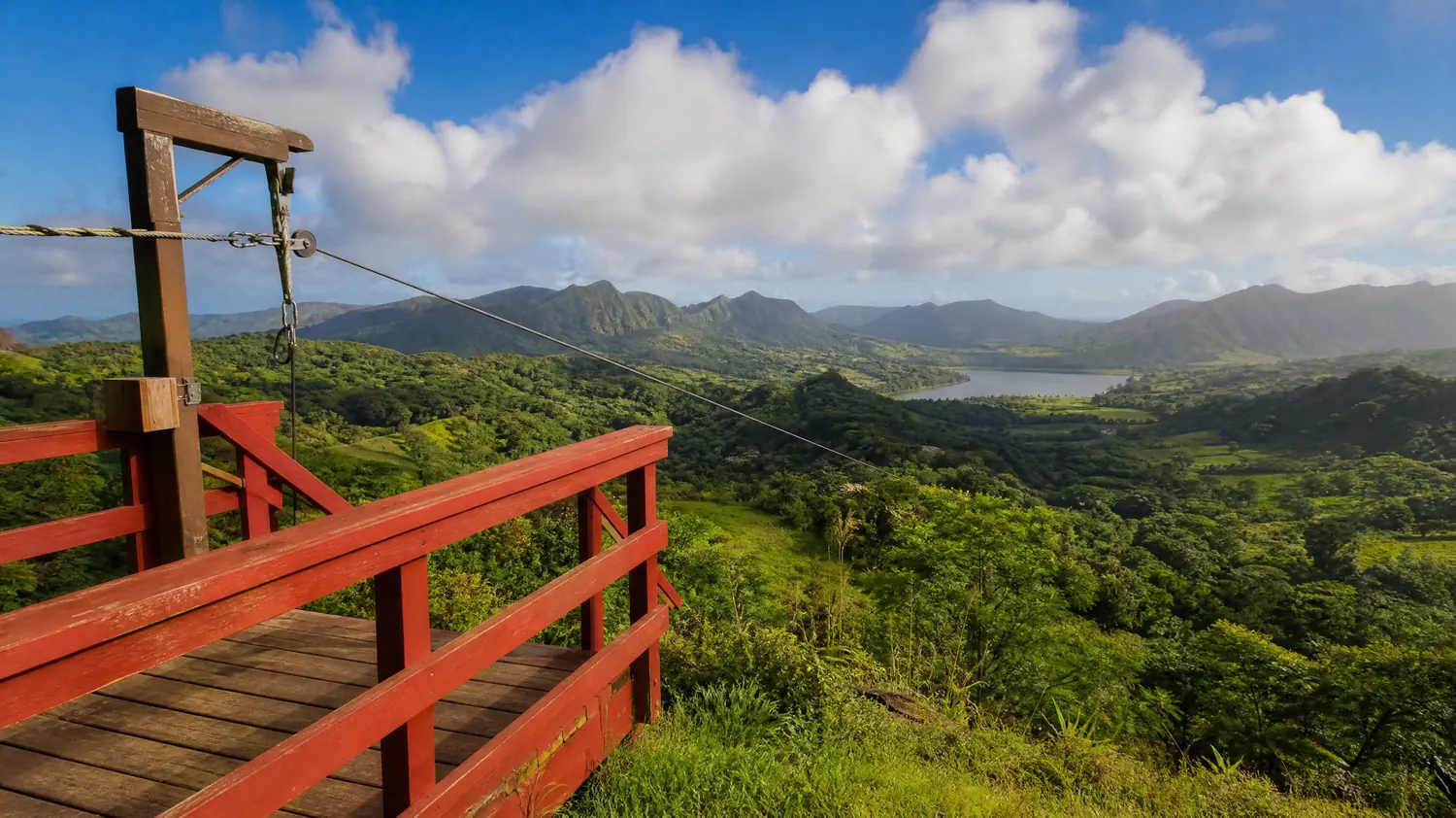 A zipline launch platform with red railings above a green valley and a distant reservoir